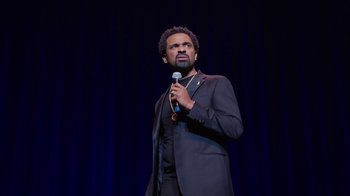 Movie still from “Mike Epps: Only One Mike” (2019), directed by Kevin Bray – A man in a black suit holding a microphone; Medium shot, Over the shoulder angle