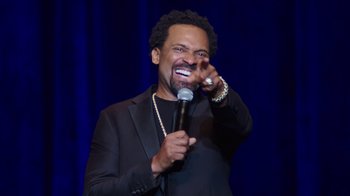 Movie still from “Mike Epps: Only One Mike” (2019), directed by Kevin Bray – A man holding a microphone in front of a microphone stand; Close Up shot, Over the shoulder angle