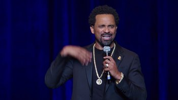 Movie still from “Mike Epps: Only One Mike” (2019), directed by Kevin Bray – A man in a black jacket is holding a microphone; Close Up shot, Over the shoulder angle