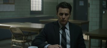 Movie still from “Mindhunter” (2017), directed by Tobias Lindholm – A man wearing a suit and tie sitting at a table in a room; Close Up shot, Over the shoulder angle