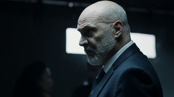 Movie still from “Money Heist” (2017), created by Álex Pina – A man with a beard and a suit and tie; Close Up shot, Over the shoulder angle