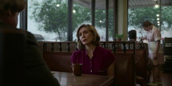 Movie still from “Love & Death” (2023), directed by Clark Johnson – A woman sitting at a table with a cup of coffee in front of a window; Medium shot, Over the shoulder angle