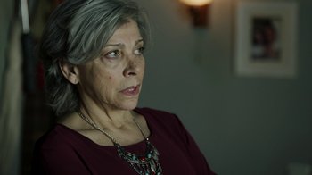 Movie still from “Money Heist” (2017), created by Álex Pina – An older woman wearing a red shirt and silver necklace; Close Up shot, Over the shoulder angle