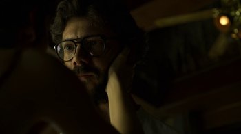 Movie still from “Money Heist” (2017), created by Álex Pina – A man with a beard and glasses is sitting in a dark room; Close Up shot, Low angle