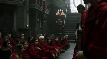 Movie still from “Money Heist” (2017), created by Álex Pina – A group of people sitting in a room with a man standing on the stairs; Wide shot, Over the shoulder angle