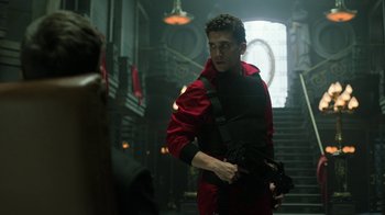 Movie still from “Money Heist” (2017), created by Álex Pina – A man in a red jacket holding a gun in front of a staircase; Medium shot, Low angle