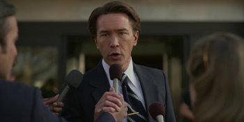Movie still from “Love & Death” (2023), directed by Clark Johnson – A man wearing a suit and tie talking into a microphone; Close Up shot, Over the shoulder angle