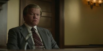 Movie still from “Love & Death” (2023), directed by Clark Johnson – A man in a suit sitting in front of a microphone; Close Up shot, Low angle