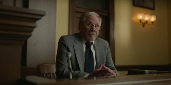 Movie still from “Love & Death” (2023), directed by Clark Johnson – An older man sitting at a table in front of a microphone; Medium shot, Low angle