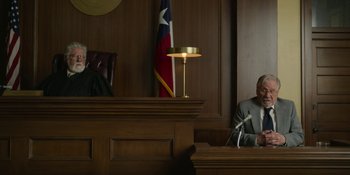 Movie still from “Love & Death” (2023), directed by Clark Johnson – A man sitting in front of a microphone in a courtroom; Wide shot, High angle