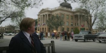 Movie still from “Love & Death” (2023), directed by Clark Johnson – A man sitting on top of a wooden bench in front of a building; Wide shot, Over the shoulder angle