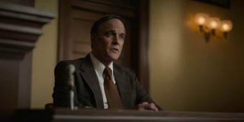 Movie still from “Love & Death” (2023), directed by Clark Johnson – A man in a suit and tie sitting in front of a microphone; Medium shot, Low angle