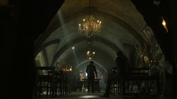 Movie still from “Money Heist” (2017), created by Álex Pina – A man standing in front of a table in a room with chandeliers; Wide shot, Low angle