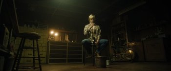 Movie still from “Monster” (2022), directed by Clement Virgo – A person sitting on a stool in a room; Wide shot, Low angle