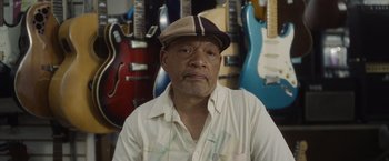 Movie still from “Monster” (2022), directed by Clement Virgo – An older man wearing a hat in front of a row of guitars; Close Up shot, Low angle