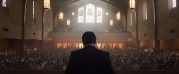 Movie still from “Monster” (2022), directed by Clement Virgo – A man in a black suit is in front of an audience; Extreme Wide shot, Low angle