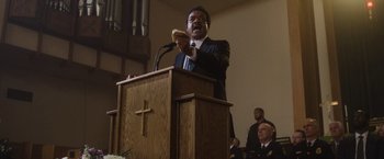 Movie still from “Monster” (2022), directed by Clement Virgo – A man in a black suit is giving a speech; Medium shot, Low angle
