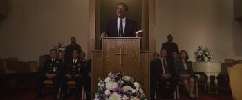 Movie still from “Monster” (2022), directed by Clement Virgo – A man in a suit is speaking at a podium; Medium shot, Low angle