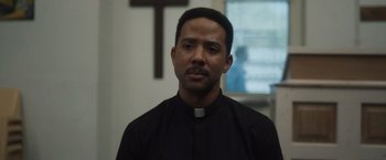 Movie still from “Monster” (2022), directed by Clement Virgo – A man wearing a priest's collar in front of a cross; Close Up shot, Low angle