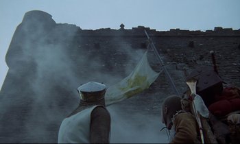 Movie still from “Monty Python and the Holy Grail” (1975), directed by Terry Gilliam – A man in a uniform holding a flag in front of a wall; Wide shot, Low angle