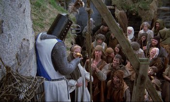 Movie still from “Monty Python and the Holy Grail” (1975), directed by Terry Gilliam – A group of people standing around a man eating a banana; Wide shot, High angle