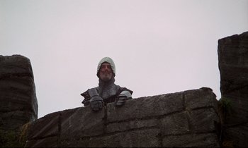 Movie still from “Monty Python and the Holy Grail” (1975), directed by Terry Gilliam – A man in a knight's outfit leaning on a stone wall; Medium shot, Low angle