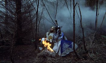 Movie still from “Monty Python and the Holy Grail” (1975), directed by Terry Gilliam – A man dressed as a medieval knight sitting on the ground by a fire; Wide shot, High angle