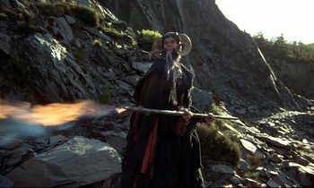 Movie still from “Monty Python and the Holy Grail” (1975), directed by Terry Gilliam – A man with a long beard holding a stick and wearing a hat; Wide shot, Low angle