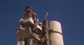 Movie still from “Life of Brian” (1979), directed by Terry Jones – A man sitting on top of a wooden structure; Wide shot, Low angle