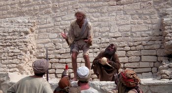 Movie still from “Life of Brian” (1979), directed by Terry Jones – A group of men in medieval garb are gathered around a stone wall; Wide shot, Low angle