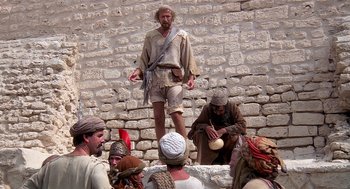 Movie still from “Life of Brian” (1979), directed by Terry Jones – A group of people sitting on a brick wall; Wide shot, Low angle
