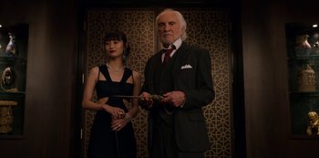 Movie still from “Murder Mystery” (2019), directed by Kyle Newacheck – An older man in a suit and a young woman in a black dress; Medium shot, Low angle
