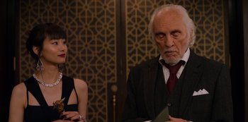 Movie still from “Murder Mystery” (2019), directed by Kyle Newacheck – An older man in a suit and a young woman in a black dress; Medium shot, Over the shoulder angle