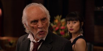 Movie still from “Murder Mystery” (2019), directed by Kyle Newacheck – An older man in a suit and tie next to an asian woman; Close Up shot, Over the shoulder angle