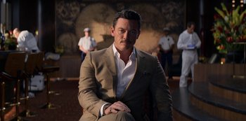 Movie still from “Murder Mystery” (2019), directed by Kyle Newacheck – A man in a suit sitting in front of a group of people; Medium shot, Low angle