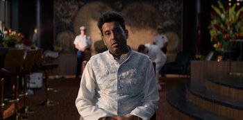 Movie still from “Murder Mystery” (2019), directed by Kyle Newacheck – A man sitting in a chair in front of other people; Medium shot, Low angle