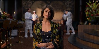 Movie still from “Murder Mystery” (2019), directed by Kyle Newacheck – A woman sitting in a chair in front of a group of people; Medium shot, Over the shoulder angle