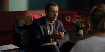 Movie still from “Murder Mystery” (2019), directed by Kyle Newacheck – A man in a suit sitting in front of a red couch; Close Up shot, Over the shoulder angle