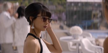 Movie still from “Murder Mystery” (2019), directed by Kyle Newacheck – A young woman wearing sunglasses and a white top; Close Up shot, Over the shoulder angle