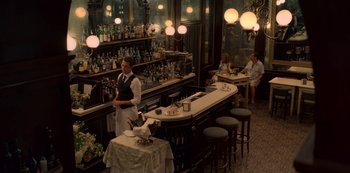 Movie still from “Murder Mystery” (2019), directed by Kyle Newacheck – A man standing at a table in front of a bar; Wide shot, High angle