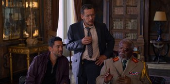 Movie still from “Murder Mystery” (2019), directed by Kyle Newacheck – A group of men sitting around each other in a room; Medium shot, Over the shoulder angle