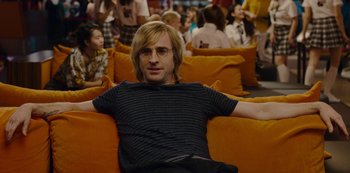 Movie still from “Mute” (2018), directed by Duncan Jones – A man sitting on top of an orange couch; Medium shot, Over the shoulder angle