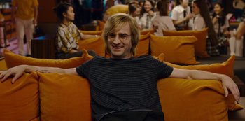 Movie still from “Mute” (2018), directed by Duncan Jones – A man sitting on a couch with his arms crossed; Medium shot, Over the shoulder angle