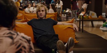 Movie still from “Mute” (2018), directed by Duncan Jones – A man sitting on top of an orange couch in a room; Medium shot, Over the shoulder angle