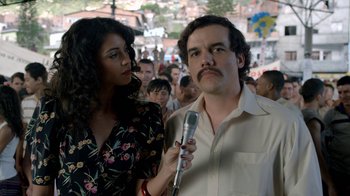 Movie still from “Narcos” (2015), created by Chris Brancato – A man and a woman holding a microphone in front of a crowd of onlookers; Close Up shot, Low angle