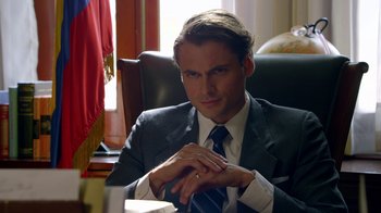 Movie still from “Narcos” (2015), created by Chris Brancato – A man in a suit and tie sitting at a desk; Close Up shot, Over the shoulder angle