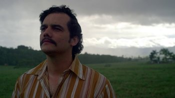 Movie still from “Narcos” (2015), created by Chris Brancato – A man in a striped shirt standing in a grassy field; Close Up shot, Low angle