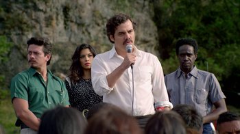 Movie still from “Narcos” (2015), created by Chris Brancato – A man is speaking to a group of people; Medium shot, Over the shoulder angle