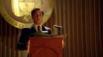 Movie still from “Narcos” (2015), created by Chris Brancato – A man standing at a podium with a microphone; Medium shot, Low angle