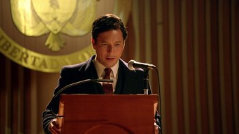 Movie still from “Narcos” (2015), created by Chris Brancato – A man standing at a podium with a microphone; Close Up shot, Low angle